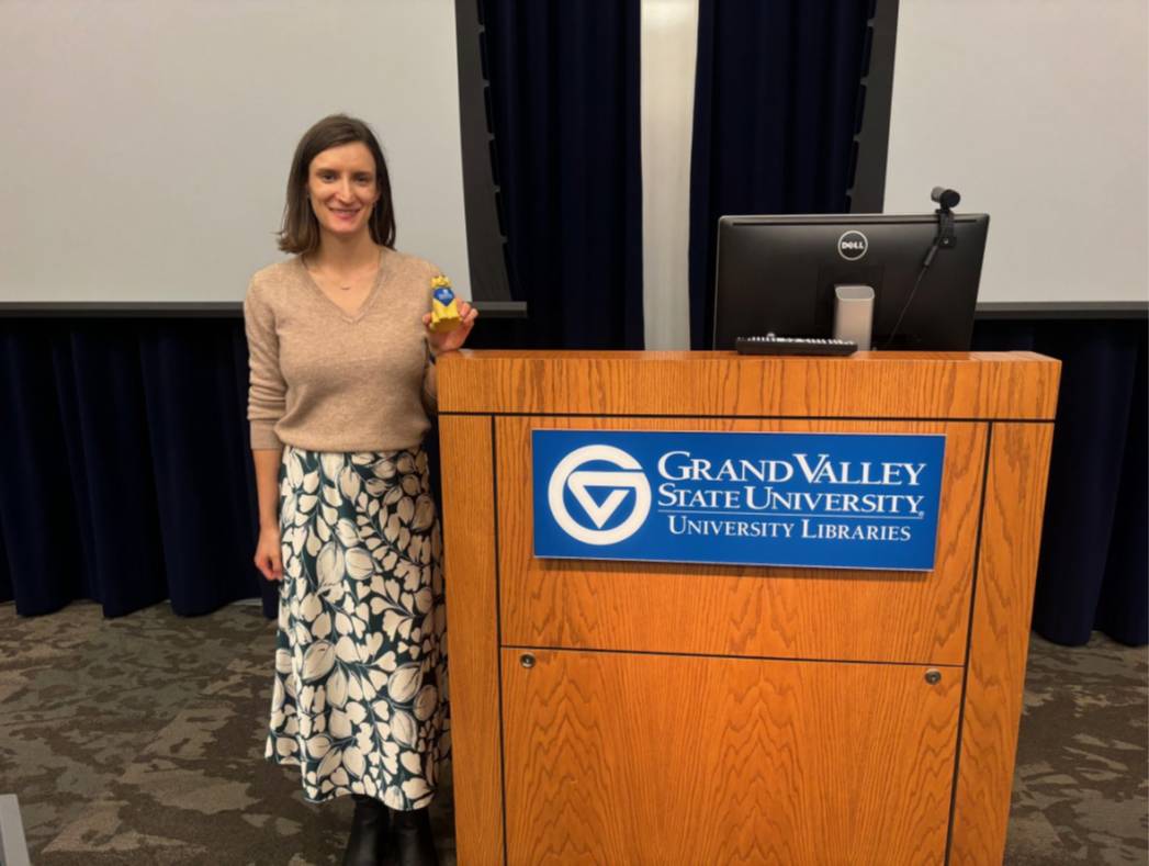 Alice Kalinowski standing next to a podium holding a GVSU History Department dog mascot toy